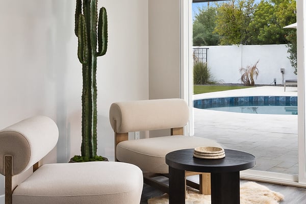 Modern living room with cream chairs, tall cactus, black table, and glass doors opening to pool and patio