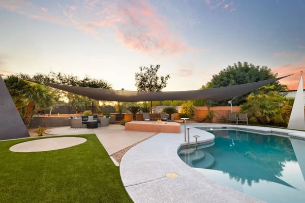Modern backyard with curved pool, lounge seating, fire pit, palm trees, and dark shade sail at sunset