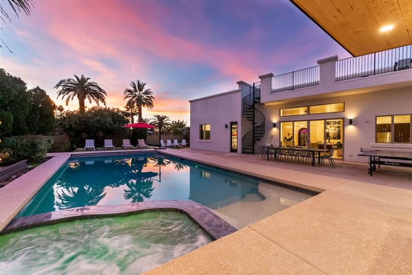 Modern house with swimming pool and spa at dusk, palm trees, lounge chairs, and lit interior at sunset