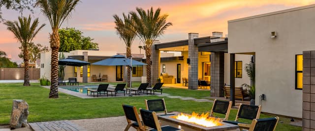 Modern home exterior at dusk with fire pit seating, pool, palm trees, and illuminated interior spaces