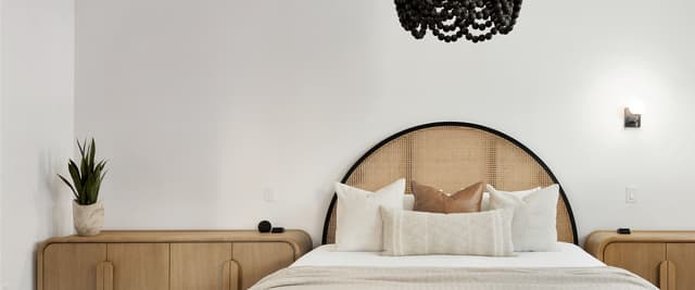 Bedroom with rattan headboard bed, cream bedding, wooden nightstands, potted plant, and black beaded ceiling fixture