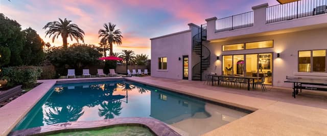Modern house with swimming pool and spa at dusk, palm trees, lounge chairs, and lit interior visible through glass doors