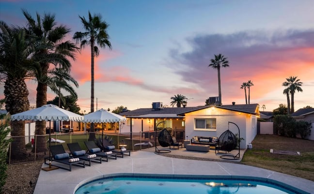 Backyard with illuminated mid-century home, pool, lounge chairs, white tent, and palm trees at sunset