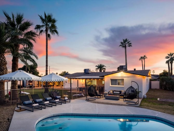 Backyard with illuminated mid-century home, pool, lounge chairs, white tent, and palm trees at sunset