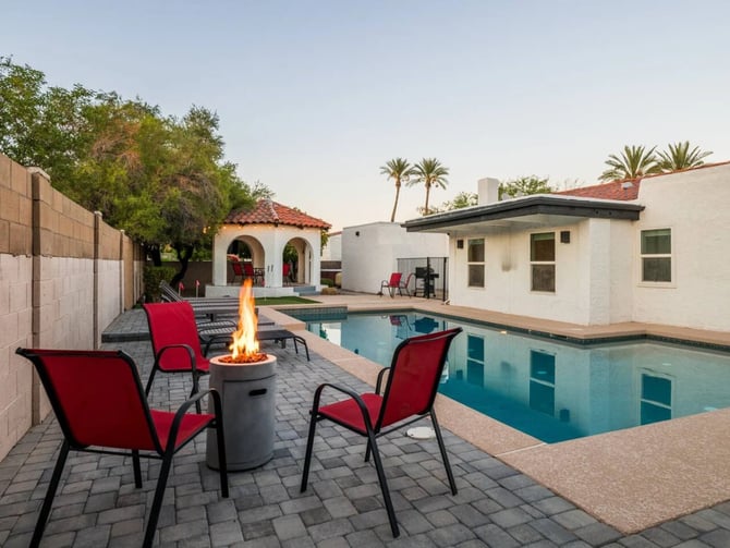 Backyard with rectangular swimming pool, fire pit with red chairs, white modern house, and Spanish-style casita with arched entryway